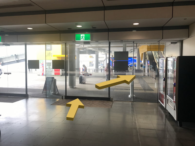 Glass doors at the airport with vending machines on the right hand side, and an escalator in the distance.