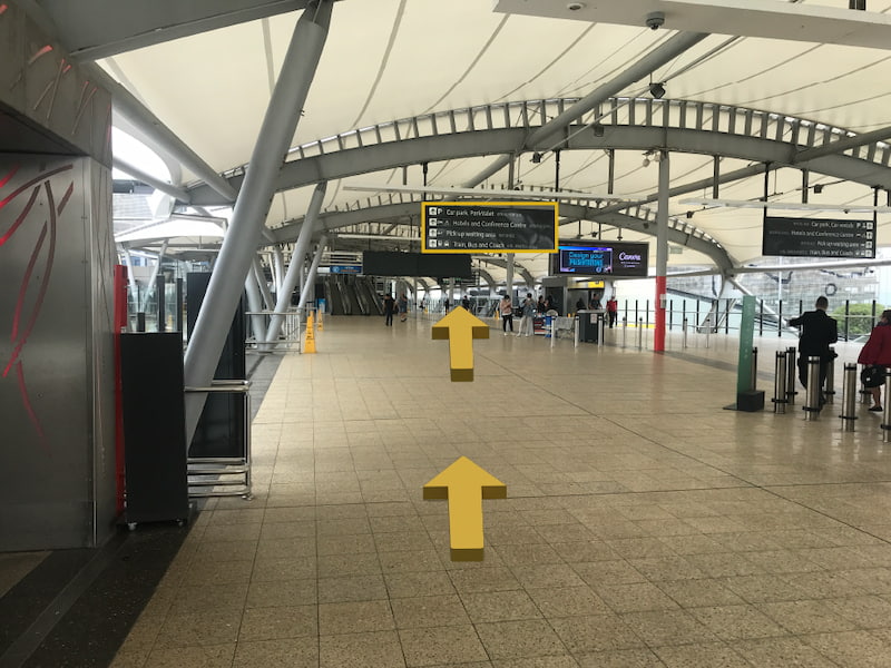 Outdoor skywalk with white roofing, people in the distance and directional airport signs.
